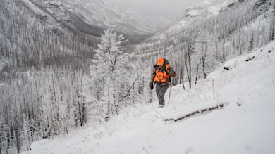 Hunter climbing a snow-covered mountainside in a valley wearing orange safety vest and cap
