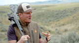 Hunter with camo rifle on shoulder wearing First Lite vest in sagebrush landscape