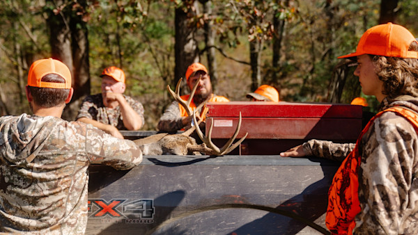 Hunters in orange caps gathered around pickup bed with deer antlers, truck decal "X4".