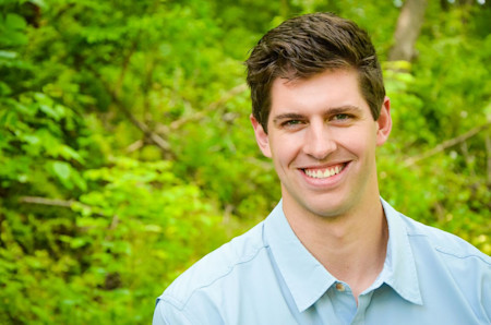 Smiling young man in light blue shirt standing outdoors against green foliage