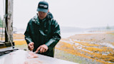 Fisherman filleting fish on metal table by shore; cap text: "can-am PRO TEAM"