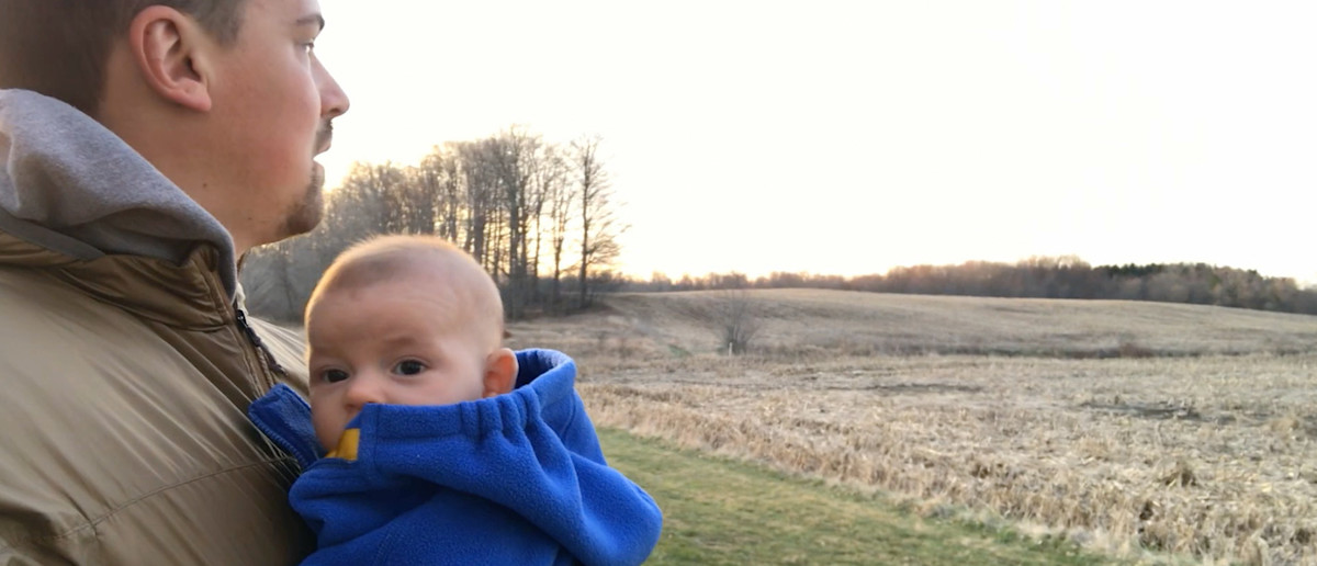 Man holding baby in blue fleece, looking across harvested field at sunset