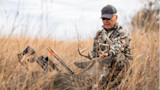 Hunter kneeling in tall grass holding buck antlers beside a crossbow; cap reads MISSION