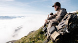 Bearded hunter sitting on alpine ridge beside packed backpack, overlooking cloud-filled valley.