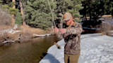 Man fishing on snowy riverbank holding rod with small fish dangling from line