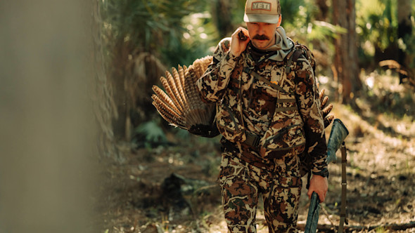 Hunter carrying turkey and shotgun in camouflage, cap reads YETI