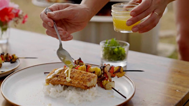 Spoon pouring yellow butter sauce over crusted fish fillet on rice beside grilled vegetable skewers