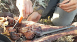Hands carving cooked game ribs with orange-handled knife and spoon outdoors