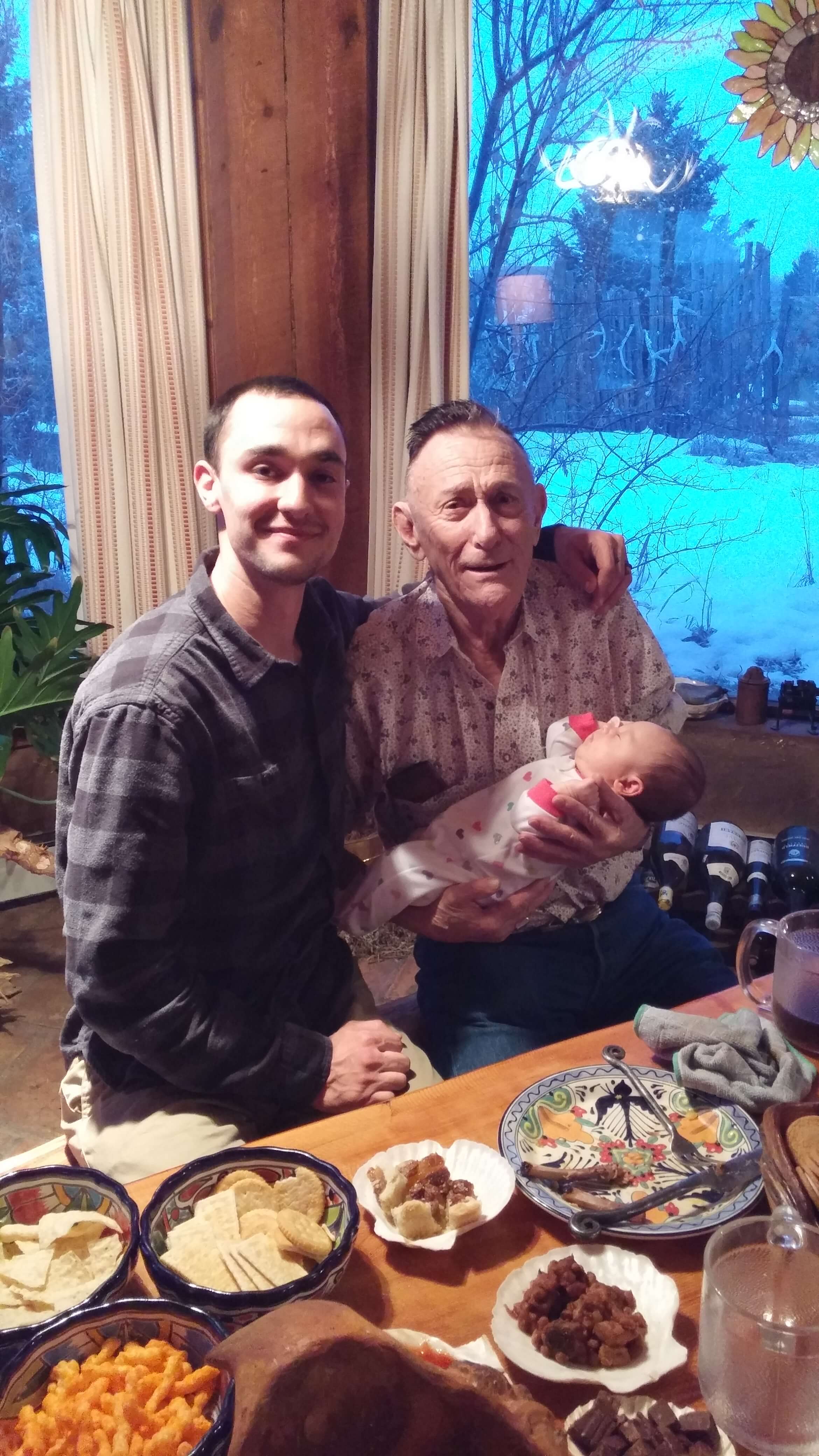 Young man with arm around elderly man holding newborn by a window with snow outside