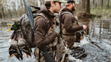 Two hunters wading in flooded woods carrying shotguns and several dead mallards
