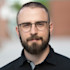 Man with round glasses and short beard looking at camera, blurred brick background