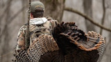 Hunter carrying wild turkey and camo shotgun in leafless woods; cap reads FIRST LITE