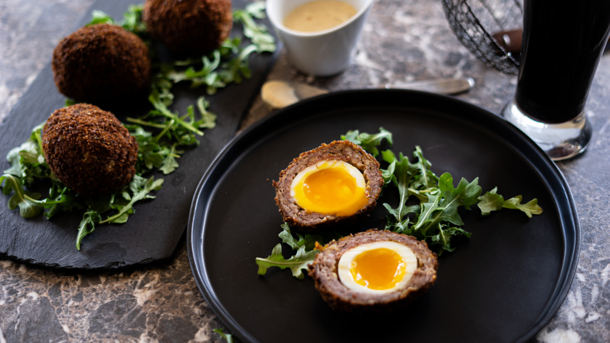 Two halved Scotch eggs with runny yolks on arugula on a black plate, whole eggs and dipping sauce nearby