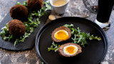 Two halved Scotch eggs with runny yolks on arugula on a black plate, whole eggs and dipping sauce nearby