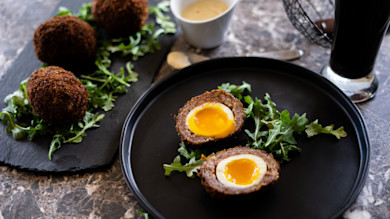 Two halved Scotch eggs with runny yolks on arugula on a black plate, whole eggs and dipping sauce nearby