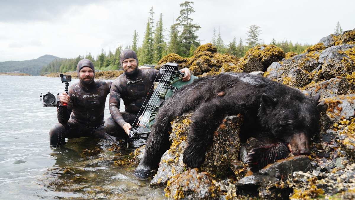 Two men in camo wetsuits with camera and bow beside dead black bear on rocky seaweed shore