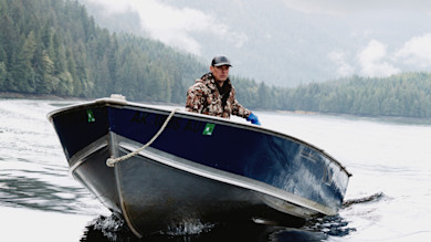 Man in camo jacket and cap steering blue boat labeled AK 1985 AU on misty forested lake
