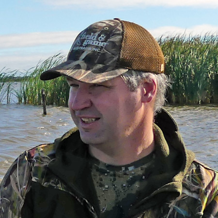 Man in camo jacket wearing cap reading "Field & Game" in shallow marsh