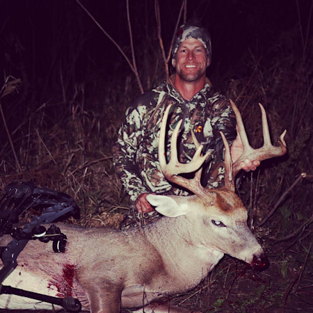 Hunter kneeling at night holding large buck antlers beside downed deer with compound bow and blood on its nose