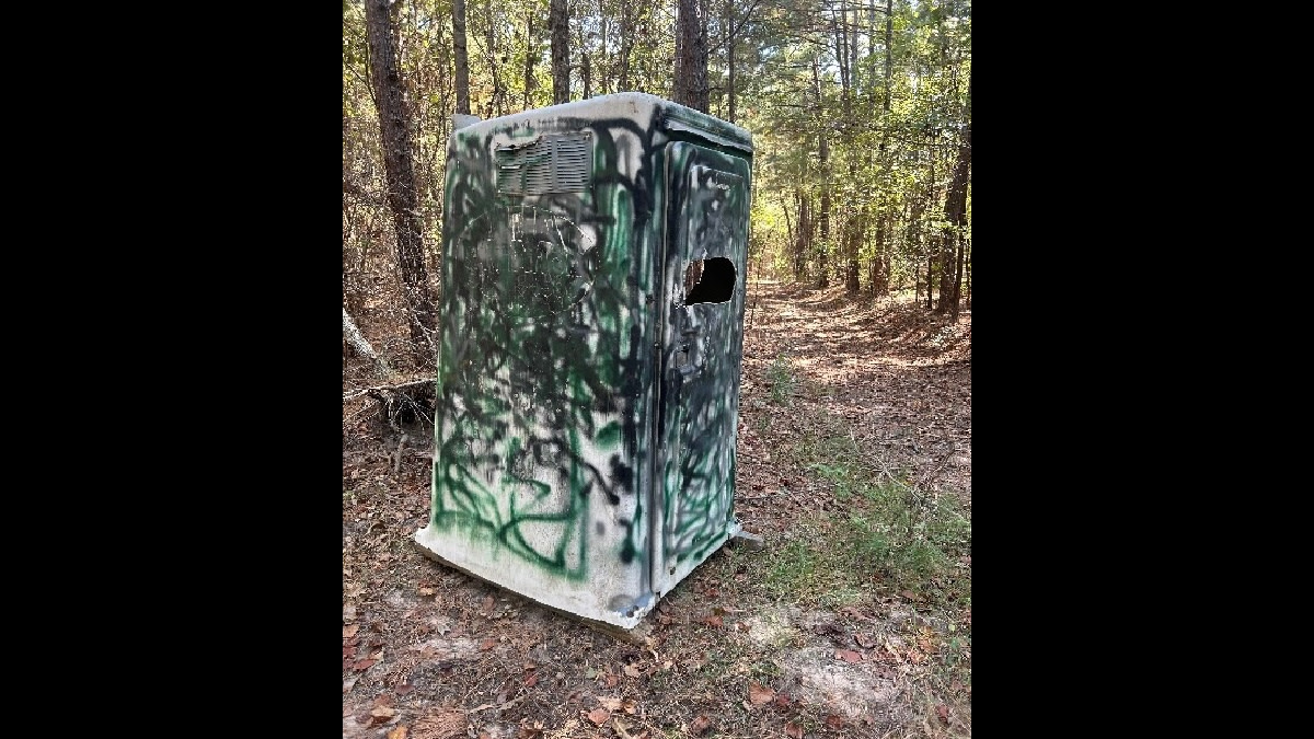 Camouflaged portable hunting blind with spray-painted graffiti and side window on forest trail
