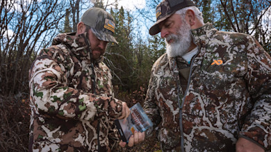 Two hunters in camo jackets and caps, one white‑bearded, holding a packet reading "AMERICAN BUFFALO"