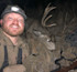 Smiling hunter wearing headlamp kneels by large buck with antlers, rifle nearby