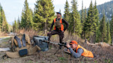 Two hunters in orange vests, one prone aiming a rifle on bipod, one standing among pines