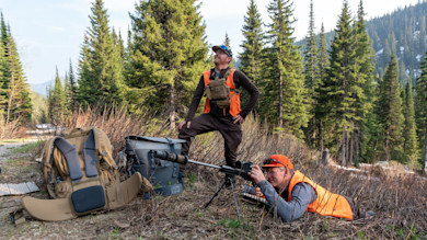 Two hunters in orange vests, one prone aiming a rifle on bipod, one standing among pines