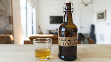 Bottle labeled "Dandelion Wine 5/9/2011" beside a glass of amber liquid on countertop