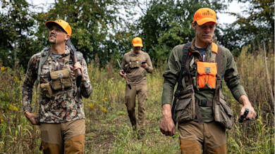 Three hunters in orange caps and camo; one carries a shotgun, one checks a phone
