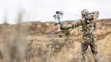 Hunter drawing a compound bow while wearing camo jacket and pants in open field
