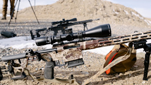 Camo precision rifle with large scope, bipod and magazine on gravel