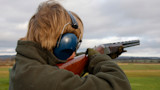 Hunter aiming over-under shotgun at open field, wearing blue earmuffs and green fleece