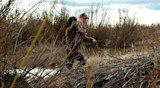 Hunter carrying beaver pelt over shoulder and holding shotgun beside creek