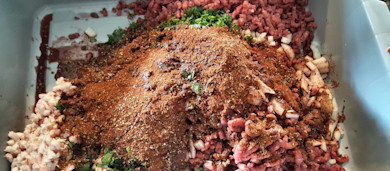Ground meat with chopped onion, fat, herbs and a mound of spices in a gray tub