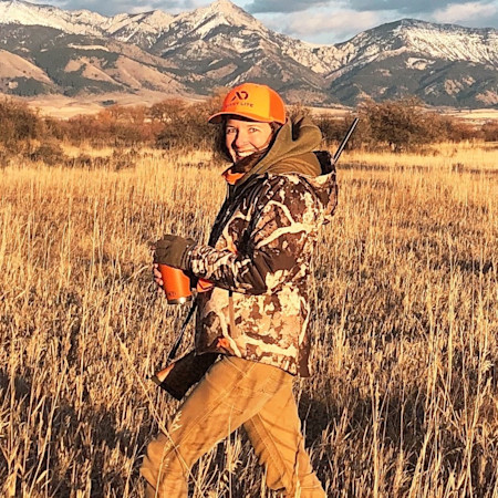 Woman hunter smiling in camouflage wearing orange First Lite cap holding cup and rifle in tall grass with mountains