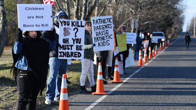 Protesters along road holding signs: "Children are Not Born to Kill" and "YOU KILL? OH, YOU MUST BE INBRED"