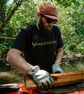 Bearded man in sunglasses wearing MEATEATER T-shirt and cap handling wood by a stream
