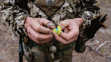 Hands holding neon yellow turkey call, hunter in FHF Gear camo jacket with tail feathers