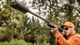 Shotgun with long suppressor aimed by hunter wearing orange cap and sunglasses in forest