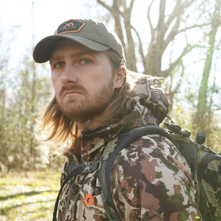 Hunter with long hair and beard wearing First Lite cap and camo jacket, backpack in sunlit woods
