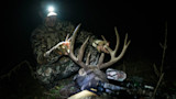 Hunter wearing headlamp holds large buck's antlers at night; compound bow in foreground