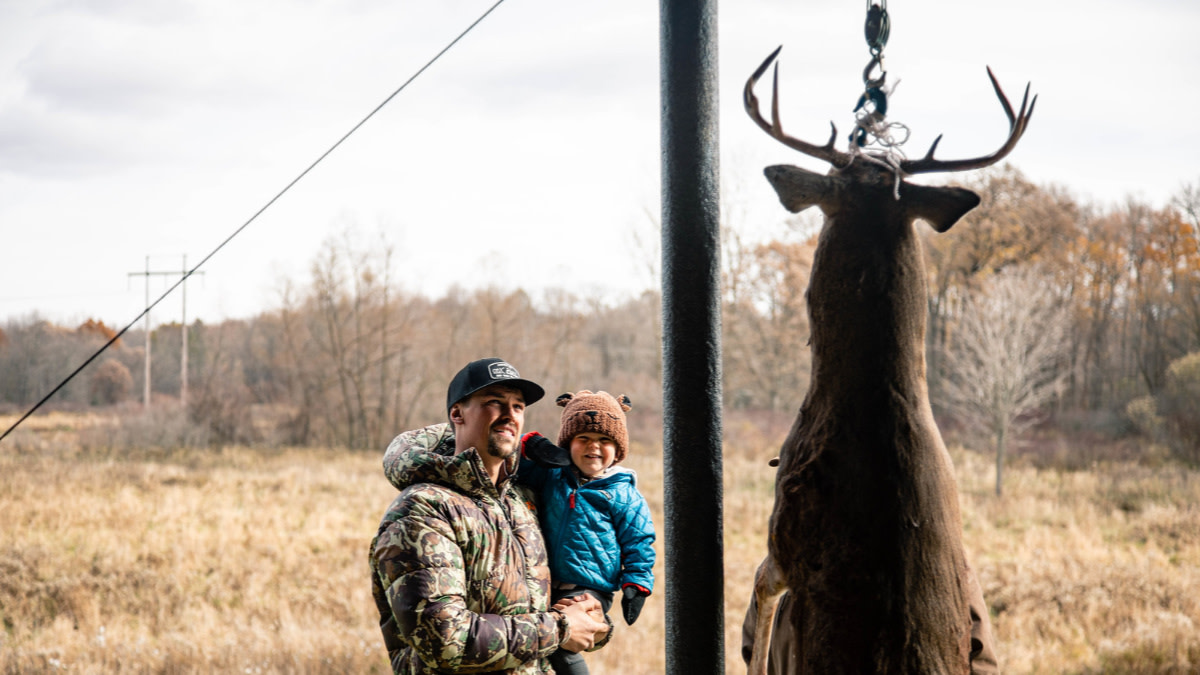 Hoisted buck beside man holding child in grassy field