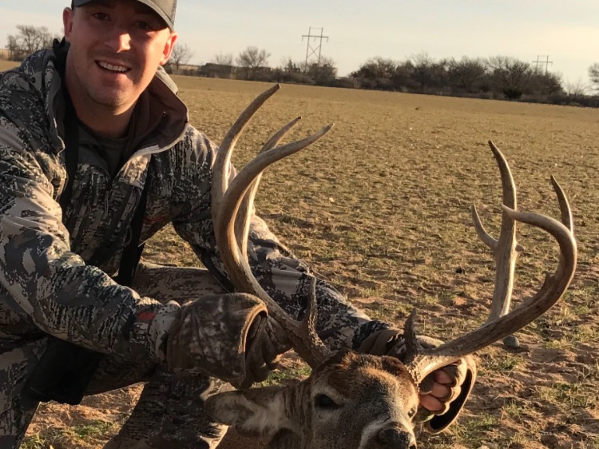 Smiling hunter kneeling in open field holding a large buck by its antlers