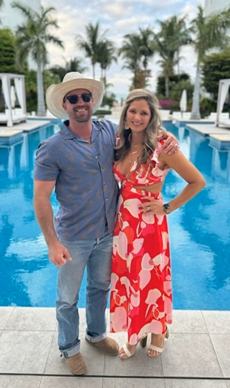 Couple smiling at a resort pool; man in straw hat and sunglasses, woman in red floral dress