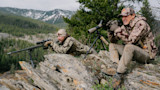 Two hunters on rocky ridge, one prone with rifle on bipod, the other seated using binoculars and a spotting scope