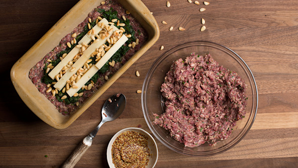 Raw wild-game meatloaf in loaf pan with spinach, cheese sticks and pine nuts; bowl of seasoned meat and mustard.
