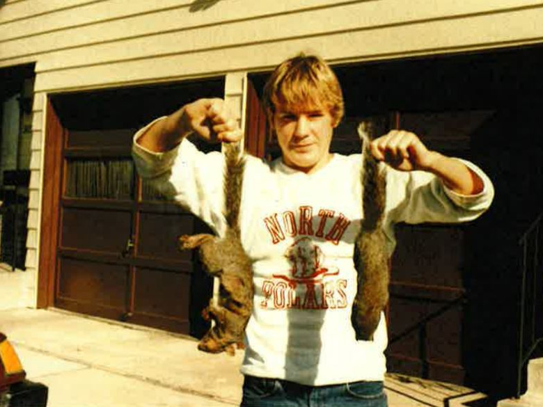 Man holding two squirrels by their tails, sweatshirt reads "NORTH POLARS", garage behind