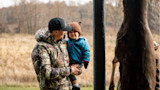 Man in camouflage jacket holding toddler in blue coat beside a hanging deer carcass in a field