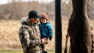 Man in camouflage jacket holding toddler in blue coat beside a hanging deer carcass in a field